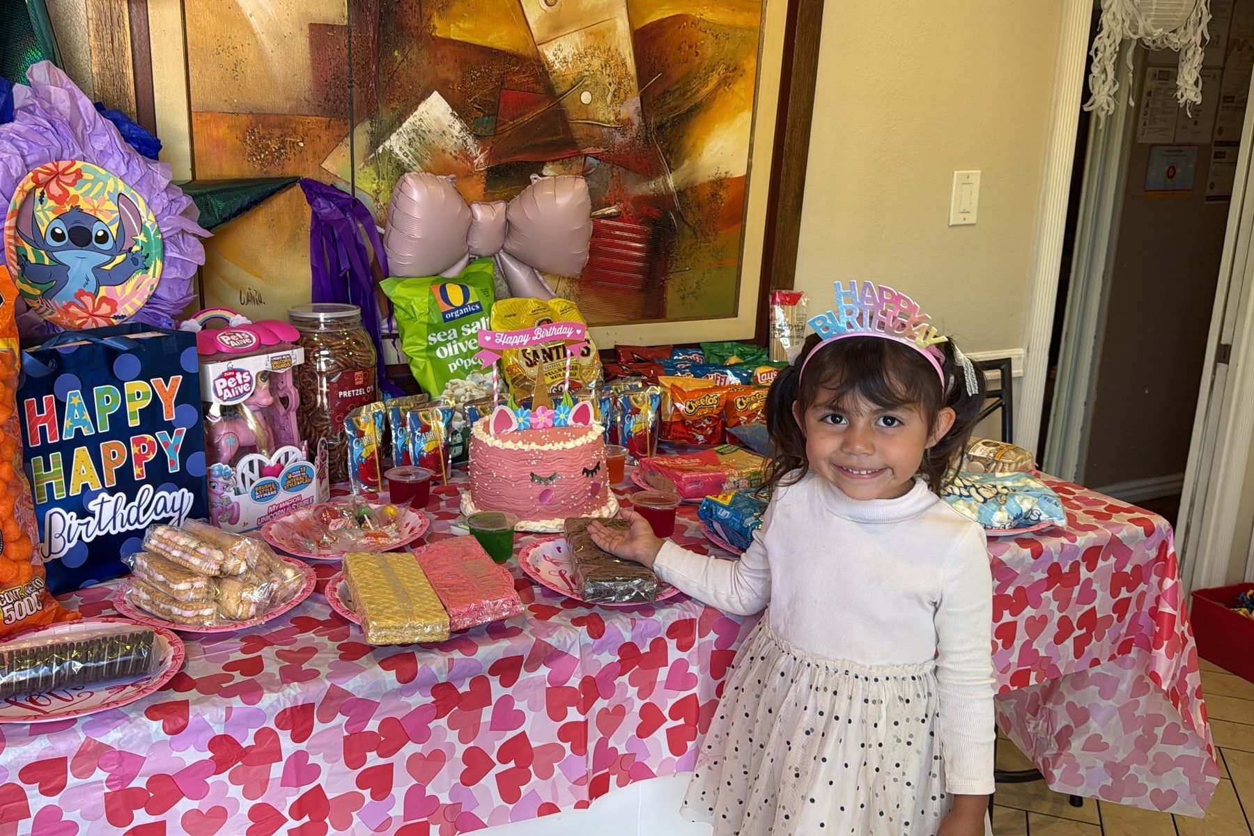 A smiling girl in a Happy Birthday crown next to her unicorn cake