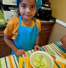 A child in a chef hat and apron slicing cucumbers